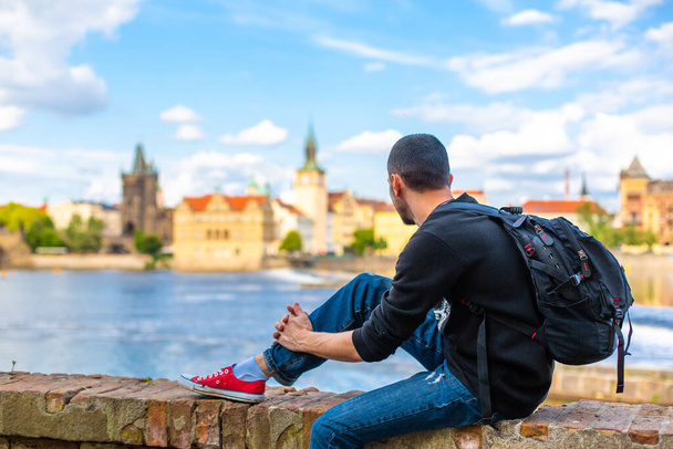 A tourist guy with a backpack sits on the embankment in Prague overlooking the Vltava river. - Foto, Imagem