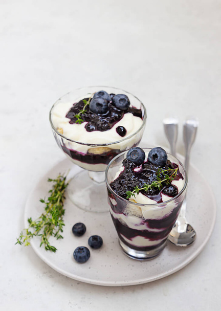 Tiramisu. Homemade dessert in glasses with blueberries, cream and ladyfingers garnish with blueberries and thyme. Light grey background. - Foto, Imagen