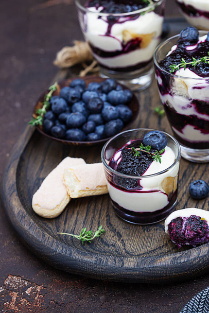 Dessert from mascarpone or ricotta cheese, blueberries and biscuit in glass on a dark background. No baked cheesecake or tiramisu. - Fotoğraf, Görsel