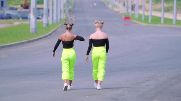 Dwie młode dziewczyny w jasnozielonych spodniach i topy z gołymi ramionami idą szeroką ulicą. Strzał z tyłu - Materiał filmowy, wideo