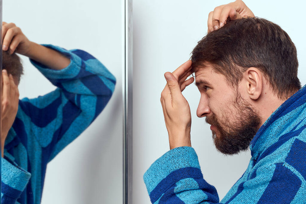 чоловік у блакитному халаті розглядає себе у дзеркалі у світлій кімнаті обрізаний вид
 - Фото, зображення
