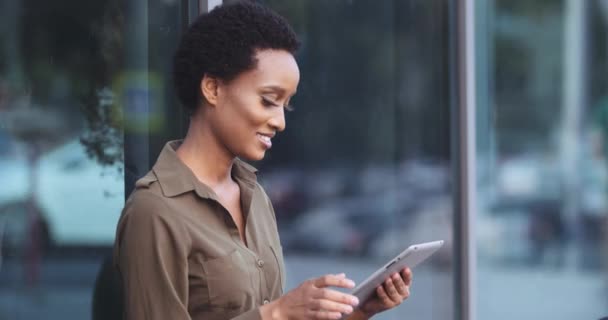 Afro-Amerikalı bir kadın cam binanın yanında oturuyor, elinde bilgisayar tableti tutuyor, internetten bilgi topluyor, parmaklarını iyi gösteren kameraya bakıyor, rıza sembolü, tamam mı? - Video, Çekim