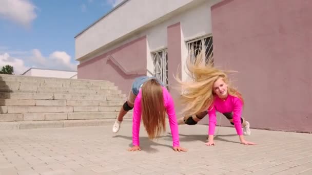 Dueto de chicas jovenes y alegres en shorts cortos bailando botines bailando delante de un edificio con una escalera. Movimiento lento - Imágenes, Vídeo