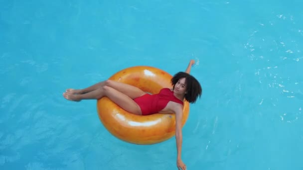 Афро-американская кудрявая модель женщина отдыхает, лежит с закрытыми глазами в оранжевом надувном спасательном буе, плавает на воде в бассейне, загорает тонким красивым телом под летним солнцем, вид сверху - Кадры, видео
