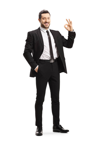 Full length portrait of a young man wearing black suit and gesturing ok sign isolated on white background - Foto, afbeelding