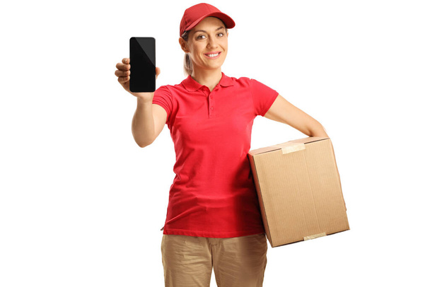 Female delivery worker in a red t-shirt holding a box and showing a mobile phone isolated on white background  - Fotó, kép