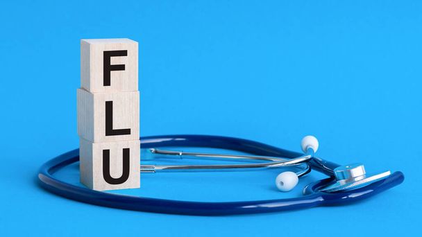 Wooden block form the word FLU with stethoscope, pills, notepad, pen on the doctor's desktop. Medical concept. Flu word written on wooden blocks and stethoscope on light blue background. insurance and medical concept - Photo, image