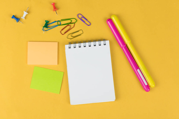 Vista superior de cuaderno en blanco y útiles escolares como marcadores de colores, pegatinas y clipers sobre fondo amarillo, espacio para texto - Foto, imagen