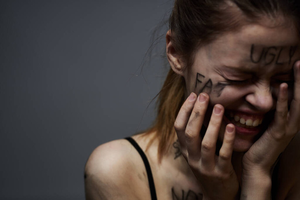 Απογοητευμένη γυναίκα με προσβλητικές επιγραφές στο σώμα της και με μαύρο μπλουζάκι σε γκρι φόντο  - Φωτογραφία, εικόνα