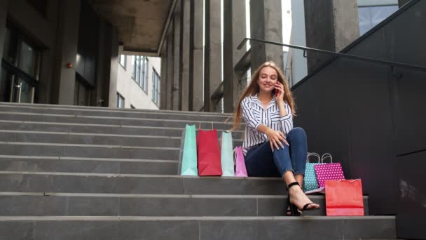 Chica sentada en las escaleras con bolsas hablando en el teléfono móvil sobre la venta en el centro comercial en el Viernes Negro - Imágenes, Vídeo