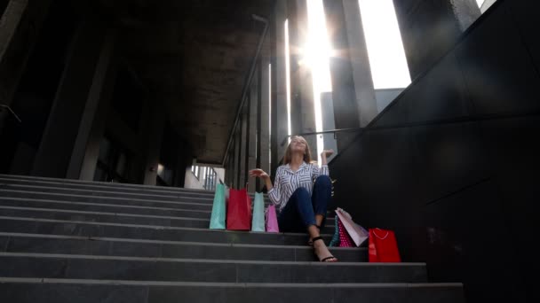 Teenager χαμογελαστό κορίτσι με τσάντες ψώνια κάθεται στις σκάλες κοντά στο εμπορικό κέντρο. Μαύρη Παρασκευή πώληση - Πλάνα, βίντεο