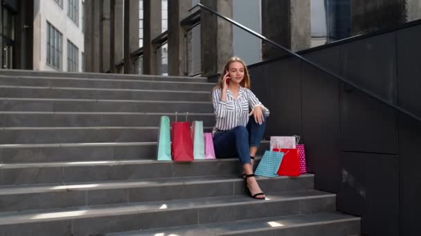 Meisje zit op de trap met tassen praten op mobiele telefoon over de verkoop in het winkelcentrum in Black Friday - Video