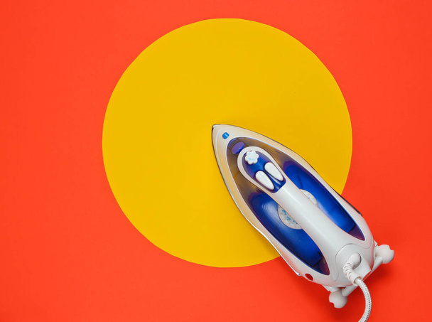 Modern iron for ironing on orange background with yellow circle in the middle. Flat lay fashion still life. Top view - Photo, Image