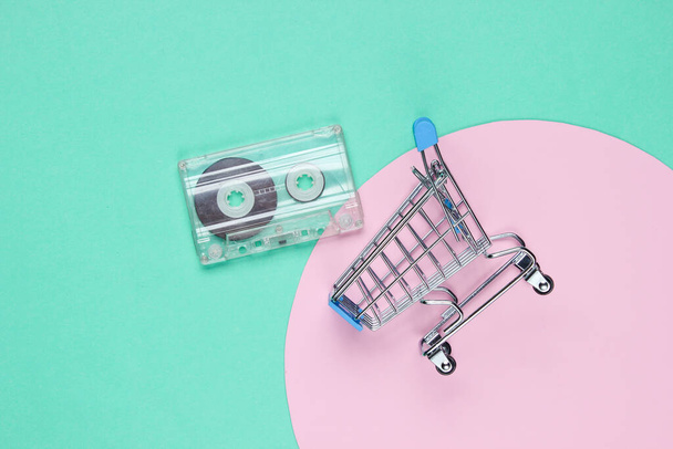 Mini carrito de compras con casete de audio retro sobre fondo azul con círculo de pastel rosa. Cultura pop. Vista superior - Foto, Imagen
