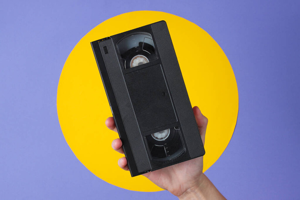 Female hand holding video cassette on purple background with yellow circle - Foto, imagen