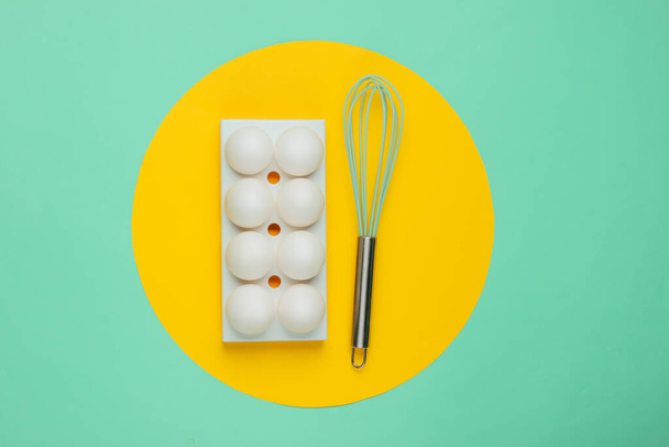 Tray with white chicken eggs and whisk on blue background with yellow circle in the middle. Minimalistic food concept. - Photo, Image