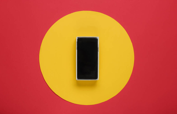 Νέο σύγχρονο smartphone σε κουτί σε κόκκινο φόντο με κίτρινο κύκλο στη μέση. Μινιμαλιστική έννοια της μόδας, δώρο - Φωτογραφία, εικόνα