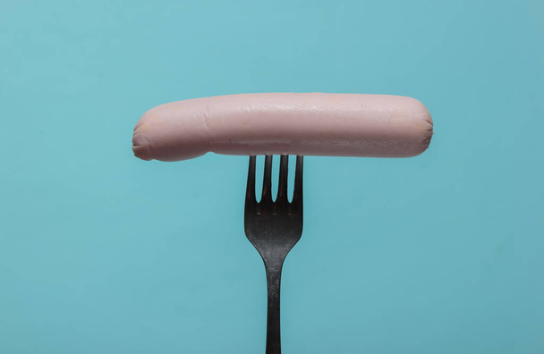 Metal fork with raw sausage on blue background. Minimalistic food concept. - Photo, Image