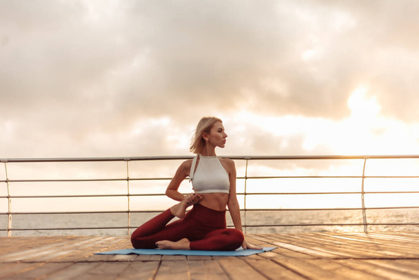 Sağlıklı bir kadın, deniz kenarında yoga yapıyor. Paspasın üzerinde oturan sporcu kadın. Eka Pada Rajakapotasana pozu - Fotoğraf, Görsel