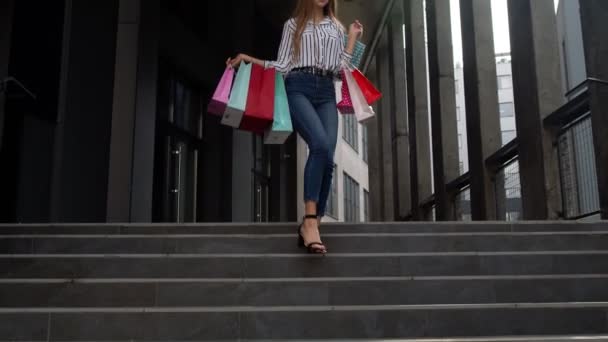Χαμογελαστό κορίτσι με τα πόδια από το κέντρο εμπορικό κέντρο με τσάντες για ψώνια, ευτυχής με χαμηλές τιμές στη Μαύρη Παρασκευή - Πλάνα, βίντεο
