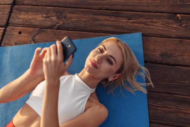Fröhliche Fitness-Frau liegt auf Matte und nutzt Smartphone im Freien - Foto, Bild
