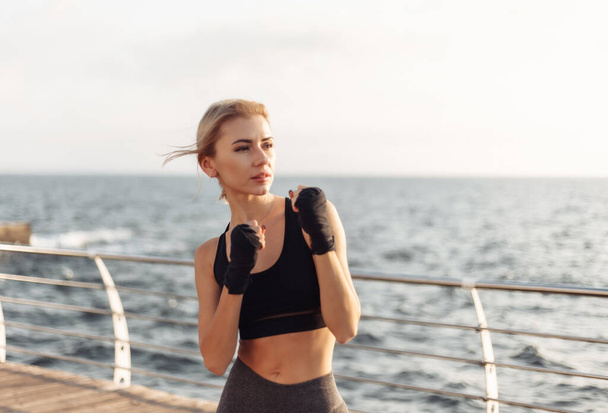 Mujer joven kickboxer en ropa deportiva con las manos envueltas en vendajes trenes en la playa al amanecer. Combate de entrenamiento matutino - Foto, imagen