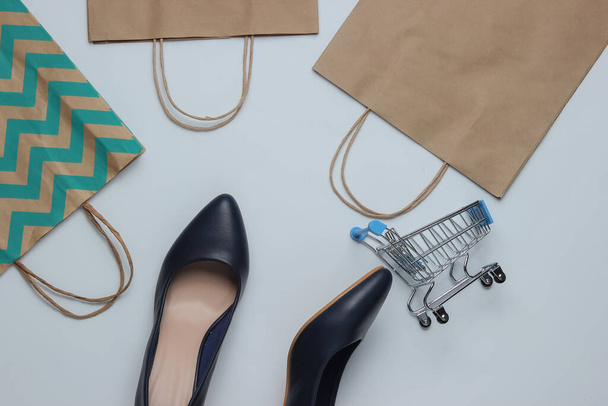 Concept de shopping. Studio de mode tourné. Chaussures à talons hauts pour femmes, chariot d'achat et sacs en papier sur fond jaune. Vue du dessus - Photo, image