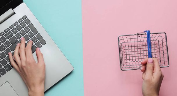 Online shopping concept. Female hands hold mini shopping basket and type on laptop keyboard on pink-blue pastel background. Top view, minimalism - Photo, Image