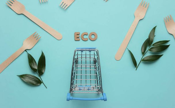Eco-vegetarian shopping. Shopping trolley, wooden forks, green leaves on blue pastel background. Top view - Photo, Image