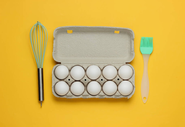 Ruoanlaiton käsite. Pahvikotelo munat, keittiövälineet (vispilä, harja) keltaisella pohjalla. Yläkuva - Valokuva, kuva