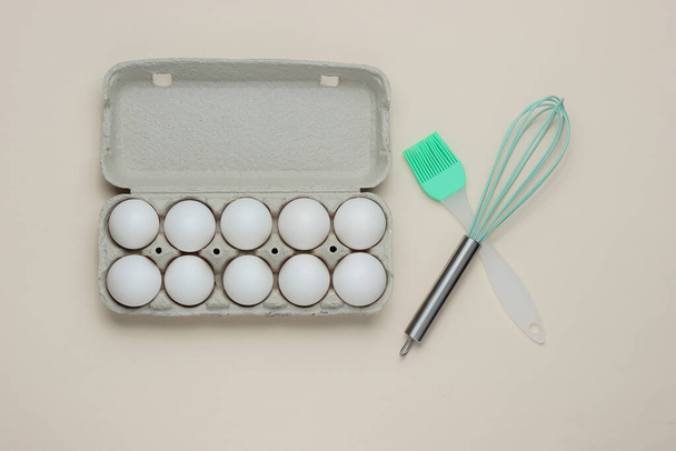 Koncept vaření. Kartónový tác s vejci, kuchyňské nářadí (šlehačka, štětec) na bílém pozadí. Horní pohled - Fotografie, Obrázek