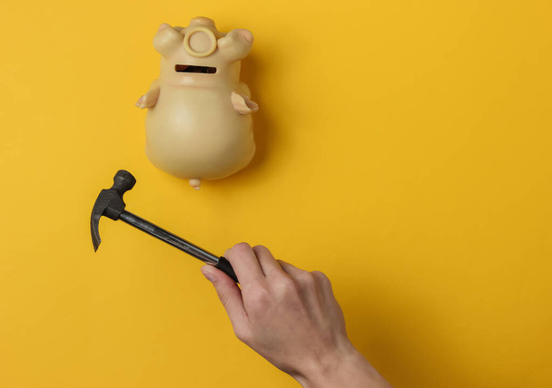 Weibliche Hand zertrümmert ein Sparschwein mit einem Hammer auf gelbem Hintergrund. Minimalistisches Konzept. Ansicht von oben - Foto, Bild