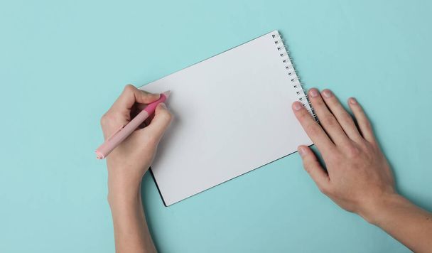 Left-handed woman writes with a pen in a diary on a blue background. Top view - Foto, immagini