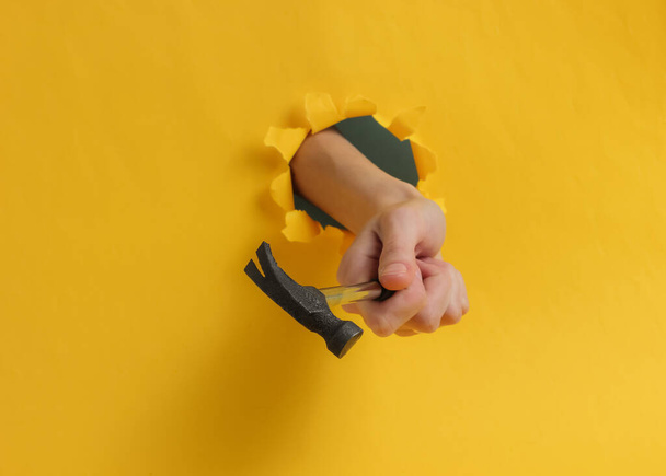 Minimalism fashion concept. Female hand holds hammer through a torn hole in yellow background - Photo, Image
