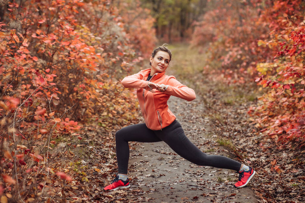 Junge attraktive Sportlerin in Sportkleidung beim Stretching im herbstlichen Wald mit geröteten Blättern von Bäumen. Aufwärmen vor dem Training. Gesunder Lebensstil - Foto, Bild