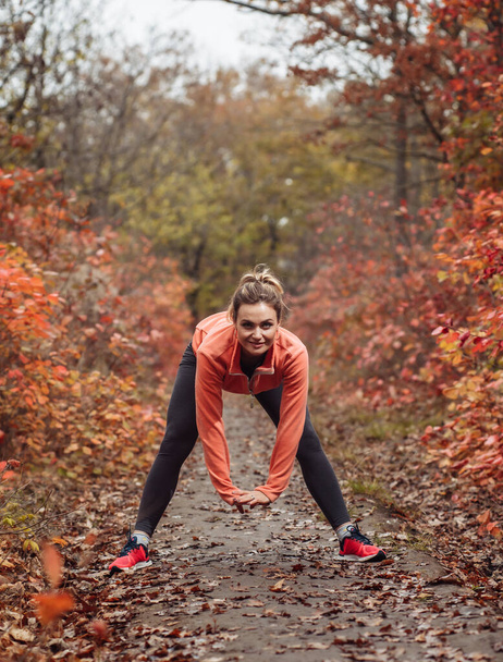 スポーツウェアの若い魅力的なスポーツ女性は、木々の緑の葉で秋の森の中でストレッチ運動を行う。訓練の前に暖まる。健康的なライフスタイルのコンセプト - 写真・画像