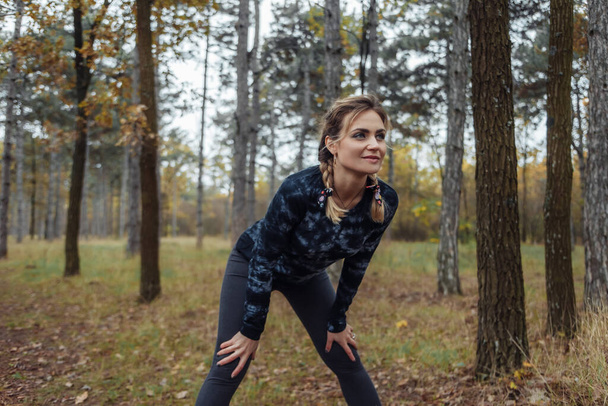 Κουρασμένος αθλητισμός ταιριάζει γυναίκα με κοτσίδες ανάπαυσης μετά το τρέξιμο στο δάσος του φθινοπώρου. Υγιεινός τρόπος ζωής - Φωτογραφία, εικόνα
