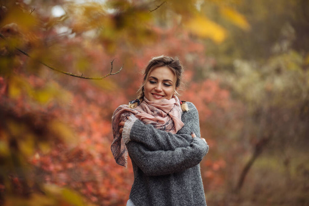 Výtvarný portrét. Mladá veselá žena v podzimním oblečení pózuje v lese s načervenalými listy stromů - Fotografie, Obrázek