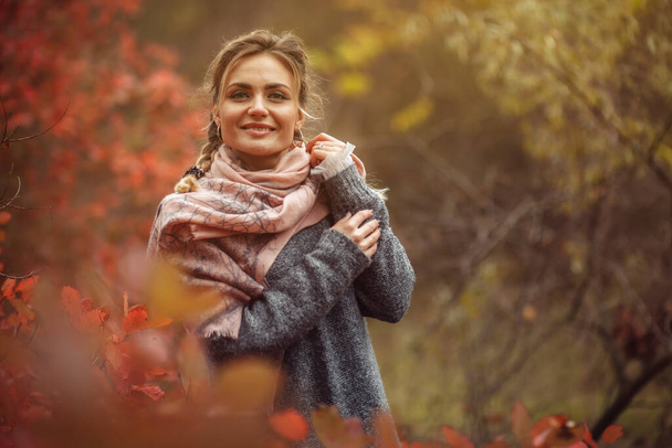 Výtvarný portrét. Mladá veselá žena v podzimním oblečení pózuje v lese s načervenalými listy stromů - Fotografie, Obrázek