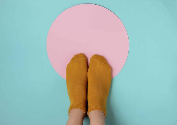 Vrouwelijke slanke poten in oranje sokken op een blauwe achtergrond met een roze cirkel in het midden. Creatief modeconcept, minimalisme. Bovenaanzicht - Foto, afbeelding