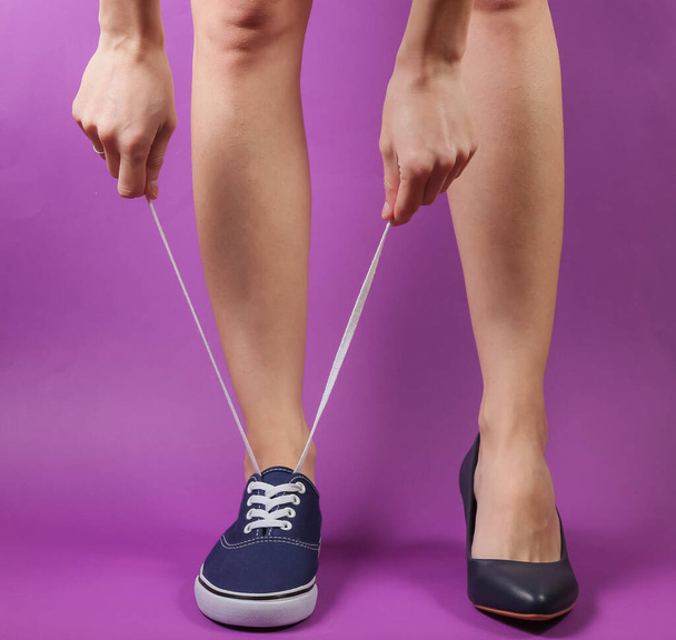 Тонкие женские ноги в двух разных туфлях на каблуках и кроссовках на фиолетовом фоне. Креативная мода - Фото, изображение