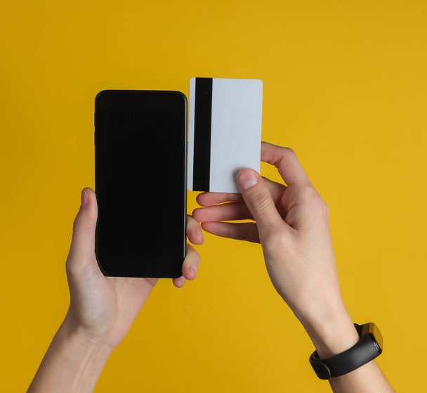 Kéz okos karkötő tartó bankkártya és okostelefon sárga alapon. Készpénz nélküli fizetés, online vásárlás. Felülnézet. - Fotó, kép