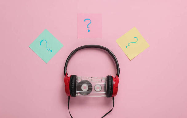 Kabelová sluchátka se zvukovou kazetou a otazníky na poznámkových kusech papíru na růžovém pastelovém pozadí. Retro styl, DJ. Top view, minimalistický hudební koncept. - Fotografie, Obrázek