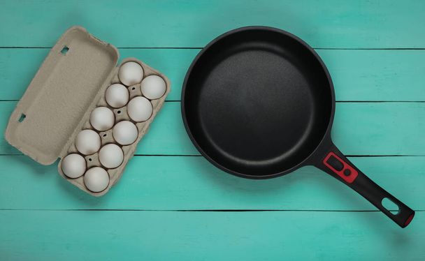Антипригарная сковорода и поднос с яйцами на синем деревянном фоне. Вид сверху - Фото, изображение