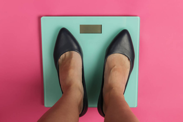 床のスケールで古典的なハイヒールの靴を持つ女性の足。ピンクのスタジオの背景。痩身コンセプト。トップ表示 - 写真・画像