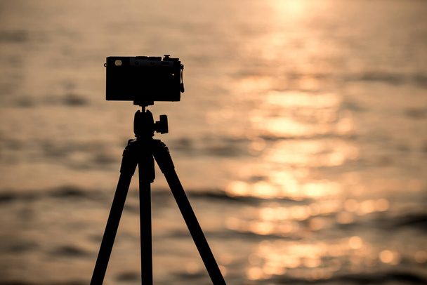 silhouette di fotocamera vintage su treppiede riprese bellissimo mare calmo con riflesso del sole sull'acqua all'alba o al tramonto - Foto, immagini