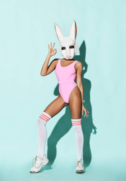 Sexy athletic slim woman model in pink swimsuit, high socks and decorative rabbit head bunny mask stands showing ok sign - Fotografie, Obrázek