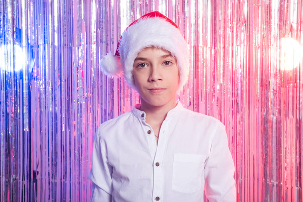 Χριστούγεννα. Εφηβικό αγόρι με το καπέλο του Αϊ Βασίλη πάνω από λαμπερό εορταστικό φόντο. Χριστουγεννιάτικες διακοπές. - Φωτογραφία, εικόνα