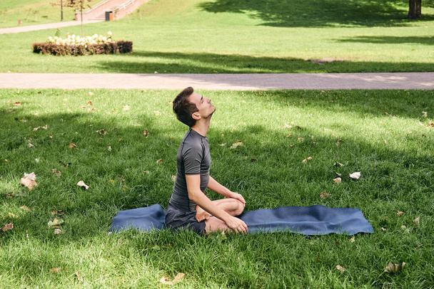 Spor kıyafetli genç adam parkta yoga yapıyor. Asana 'yı dışarıda pratik yap. Yeşil çimlerde yoga minderiyle egzersiz yapmak. Lotus pozunda oturan adam, sakin ve meditasyonlu. Fitness ve sağlıklı yaşam tarzı. - Fotoğraf, Görsel