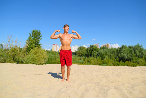 Young sporty naked man shows strength stands on the sand - Photo, Image